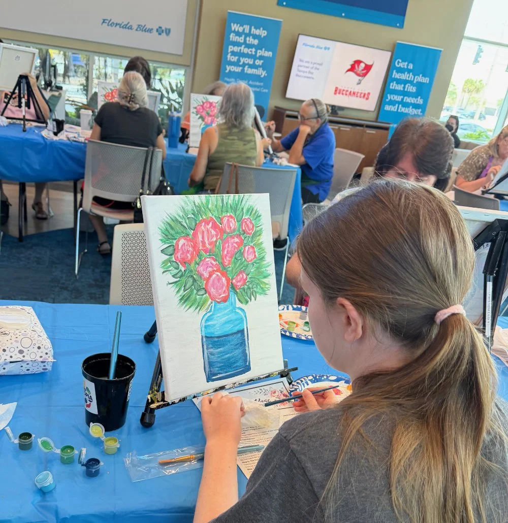 A girl painting a still life of pink flowers in a blue vase on a canvas during a group art activity in a well-lit room with other people, some painting and some working at computers, and promotional signs on the wall.