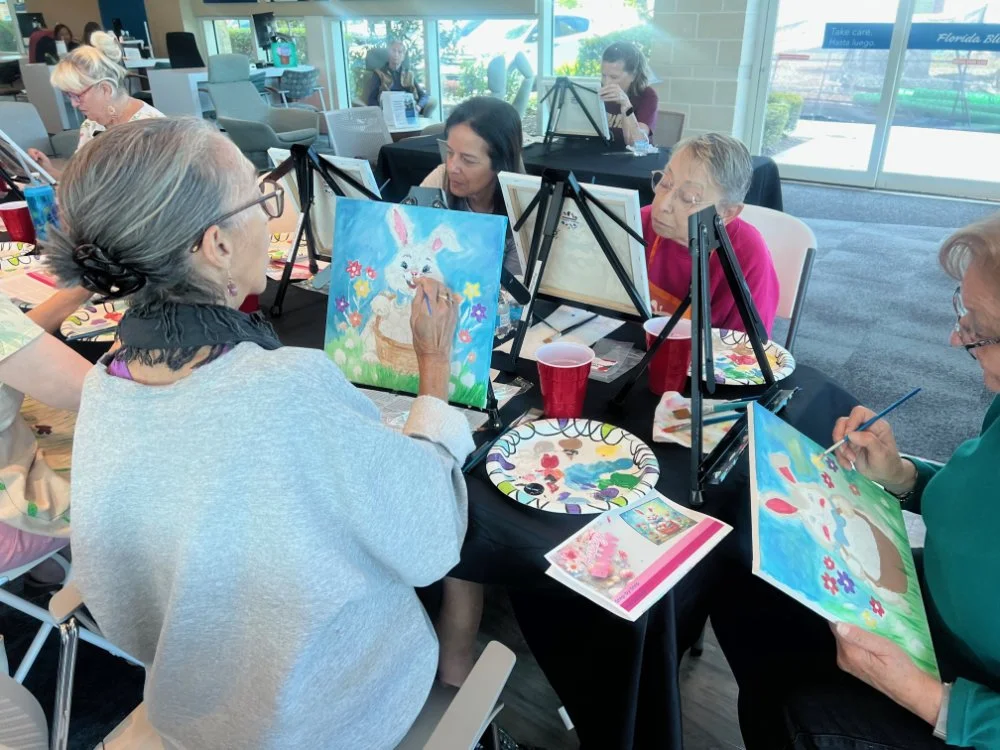 Several women painting colorful, whimsical pictures of a white bunny with flowers and a basket in an indoor setting, possibly a painting class or social gathering.