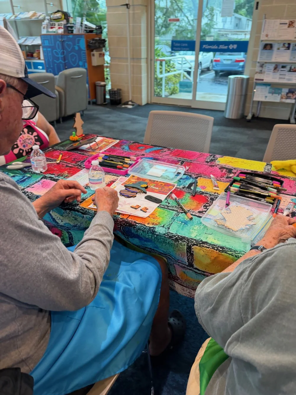 People sitting at a table covered with a colorful tablecloth, working on craft projects with markers, scissors, and foam cutouts.