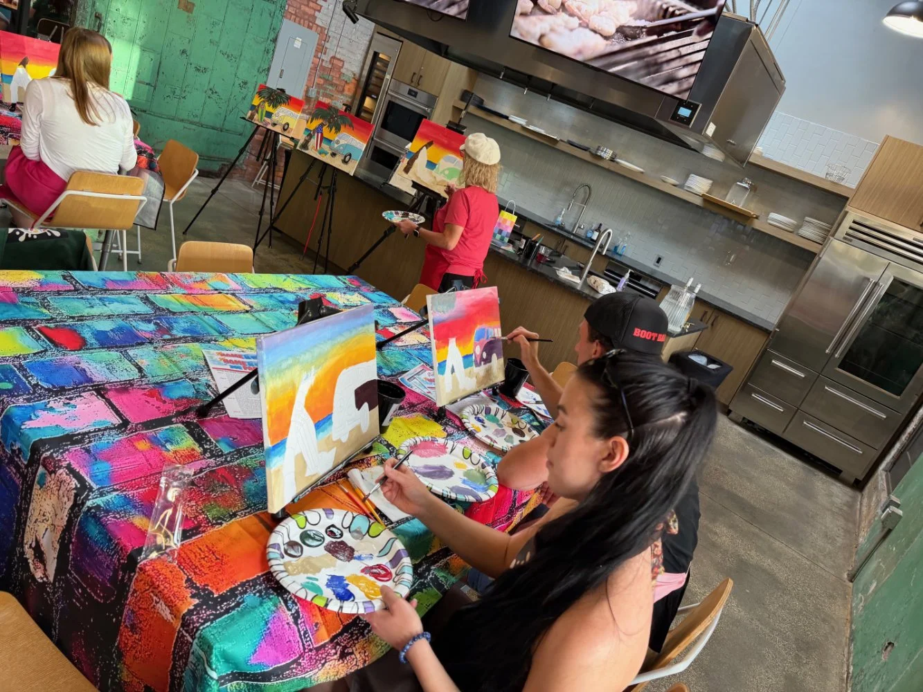 People participating in a painting class, creating colorful paintings of rainbows and dinosaurs on canvases at a table covered with vibrant, rainbow-colored cloth in a modern kitchen setting.