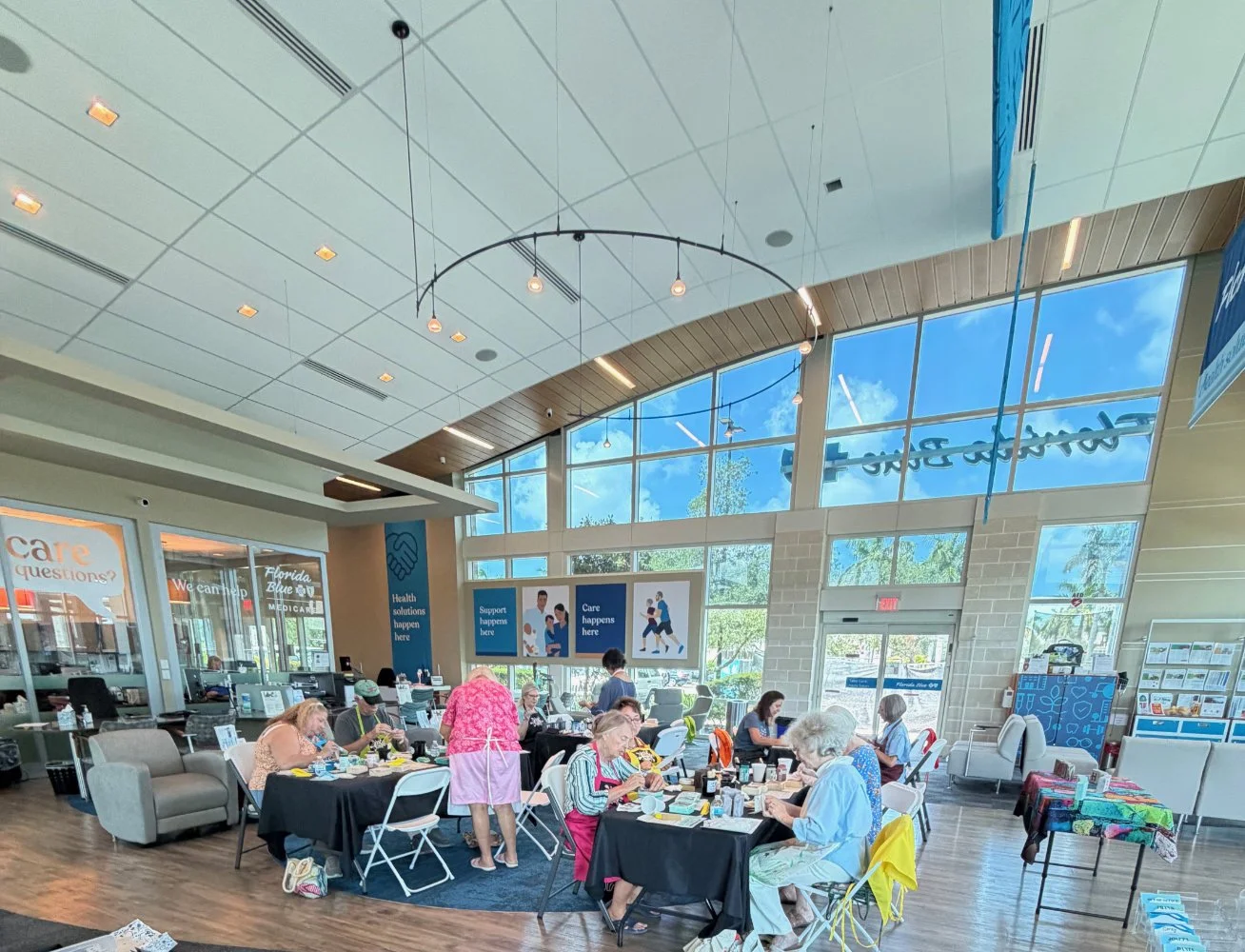 Indoor community space with several elderly women sitting at tables, engaging in activities or crafts. Large windows with a view of trees and blue sky outside, modern ceiling, and signage on the walls indicating a healthcare or community center.