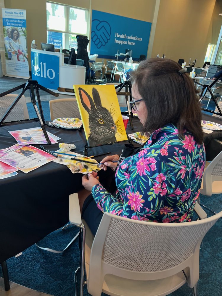 A woman with glasses and long dark hair, wearing a colorful floral jacket, is painting a black rabbit on a canvas in a bright room. The room is set up for art or community activity, with tables, chairs, and arts supplies. In the background, there are