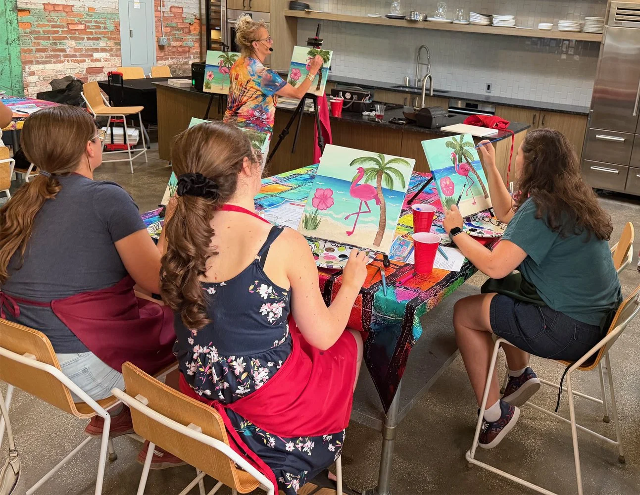 A group of women participating in a painting class in a kitchen area, with one woman painting a beach scene with pink flamingos and palm trees while the others observe, and a woman in the background demonstrating her art.