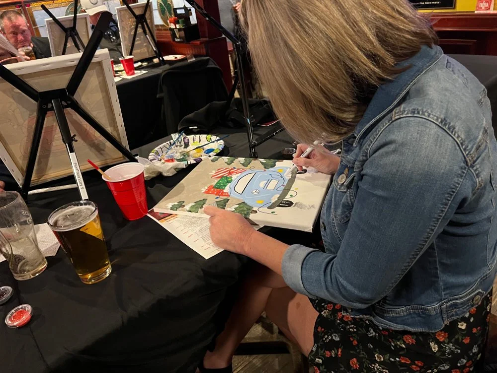 Person painting a colorful picture of a blue car with Christmas trees and gifts on a table at a social gathering.