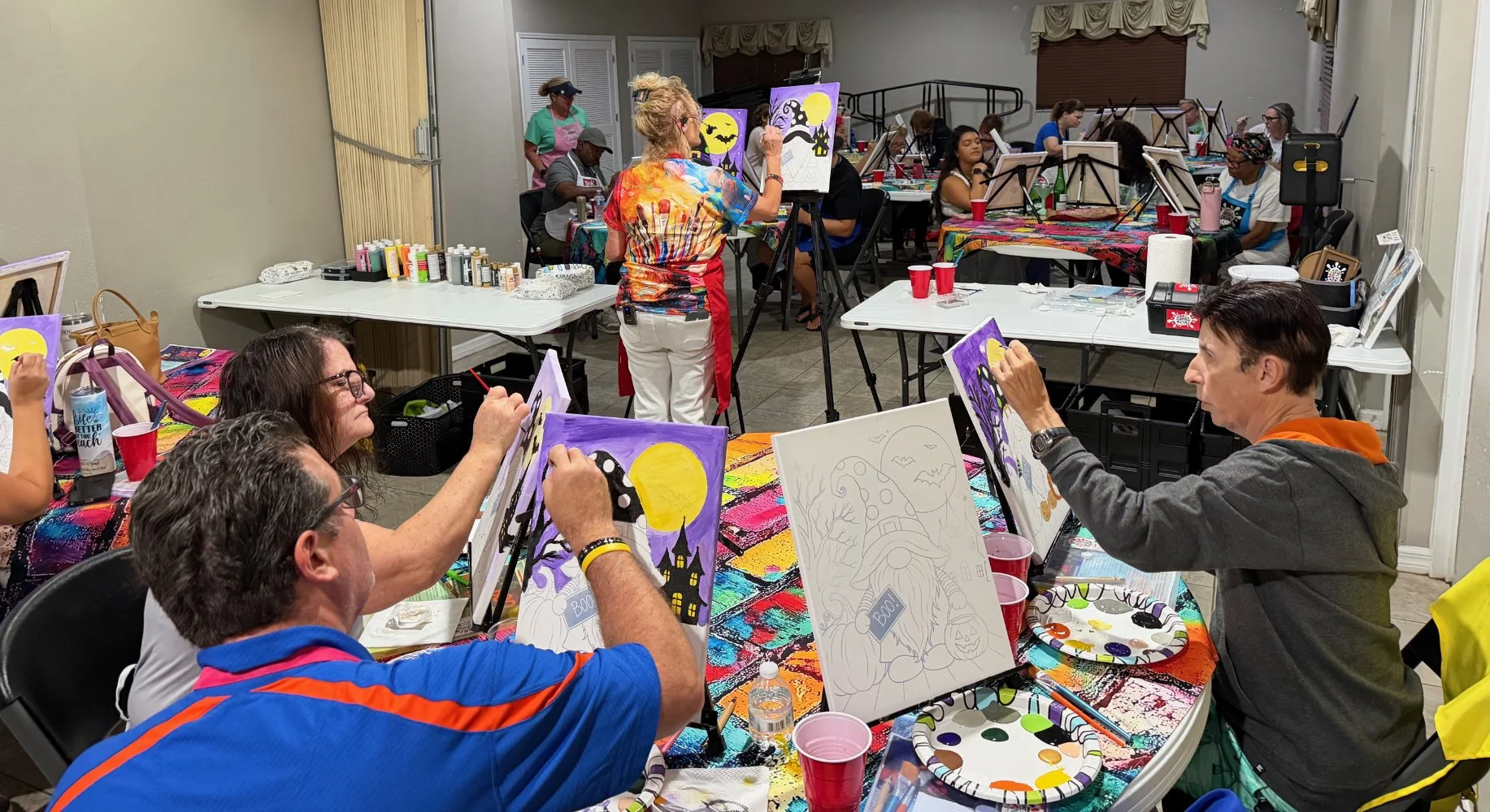 People participating in a painting class, working on Halloween-themed artwork in a decorated room, with art supplies and unfinished paintings on tables.