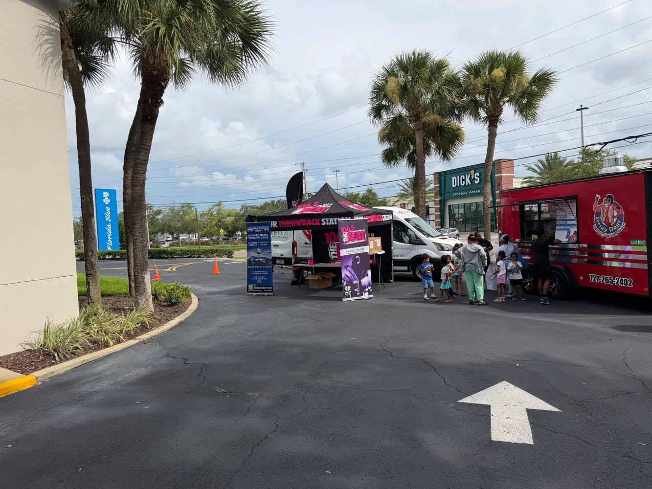 People, including children and adults, standing in line at a food truck and a promotional tent in a parking lot with tall palm trees, a Dick's Sporting Goods store, and a Florida Blue sign in the background.
