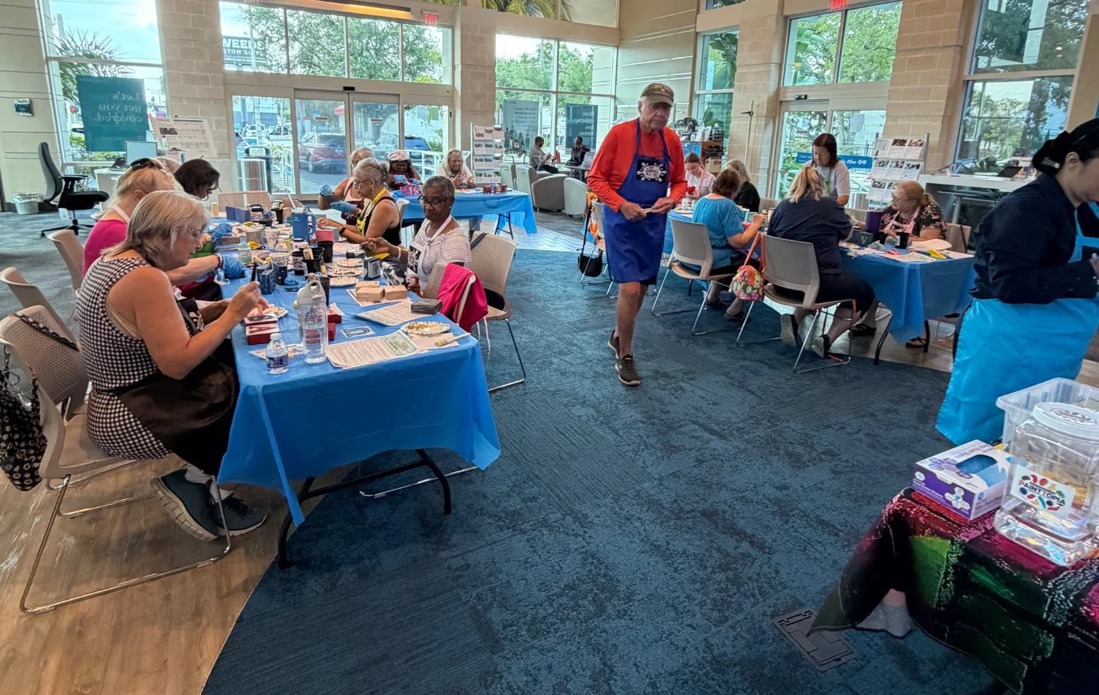 People sit around tables covered with blue tablecloths, engaged in arts and crafts, inside a bright, modern building with large windows. A man in a red shirt, blue apron, and cap walks through the room. There are storefronts and parked cars outside v