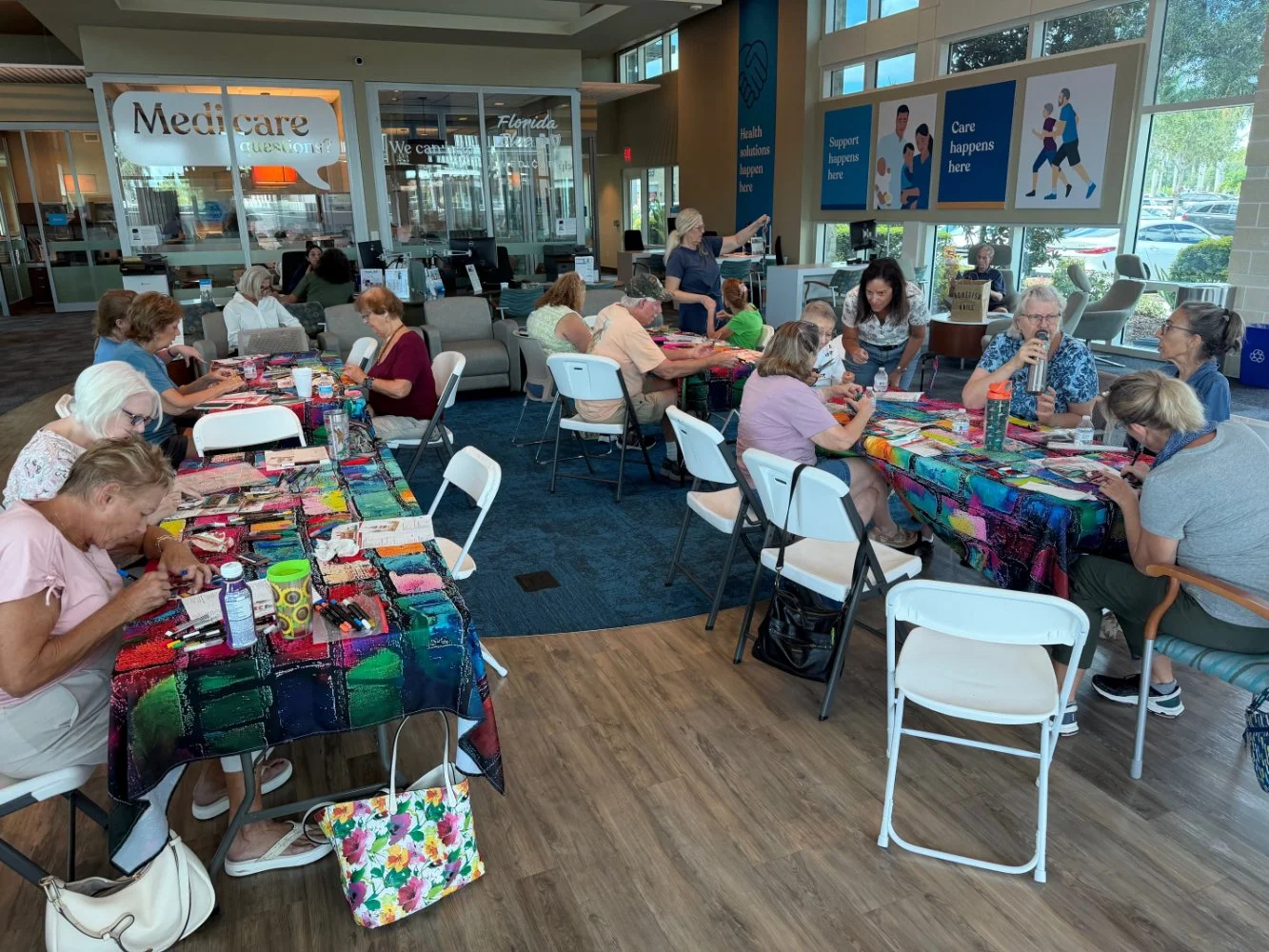 Multiple seniors participating in a craft workshop at a community center, seated at tables covered with colorful cloths and craft supplies, with windows and informational posters on the walls.