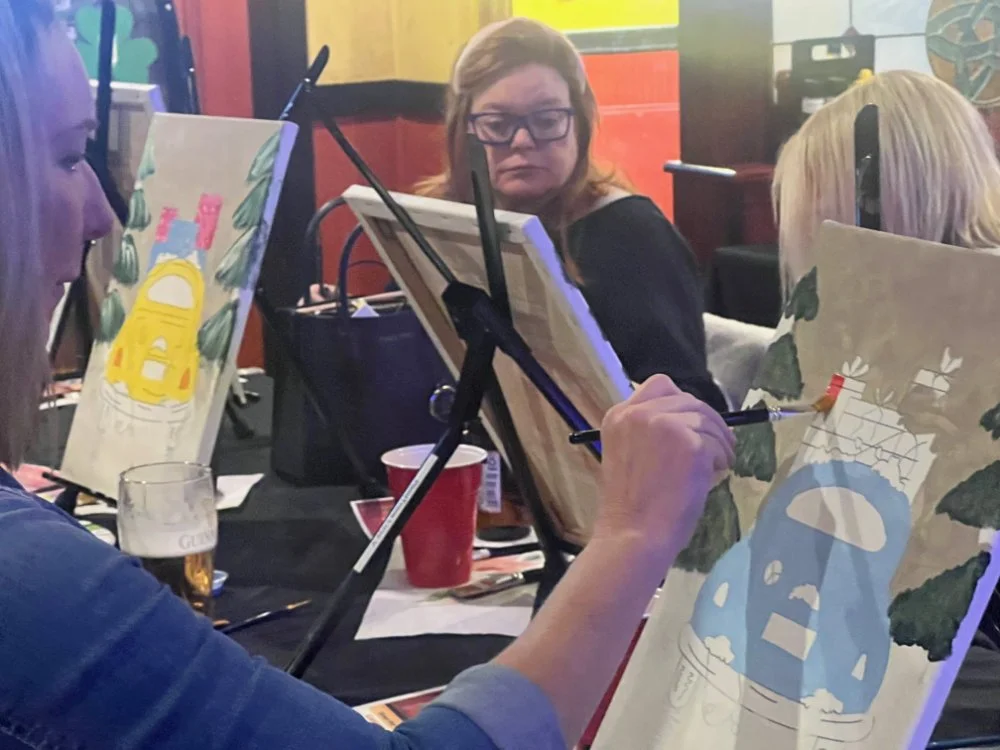 Two women painting on canvases in a lively indoor setting, possibly a bar or restaurant, with colorful decor and paintings.