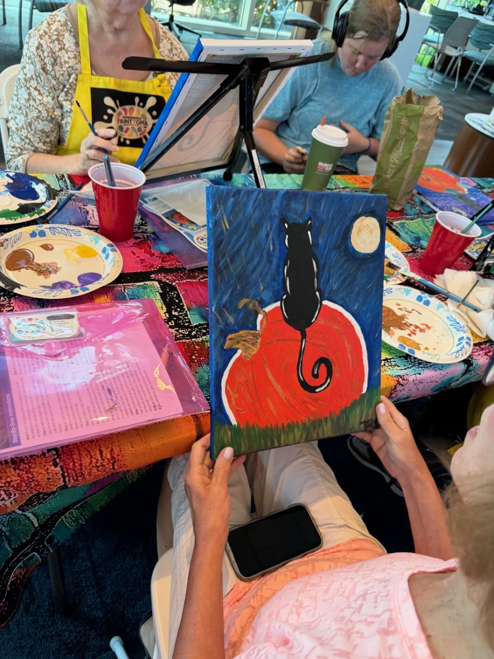 A woman holding a painting of a black cat sitting on a pumpkin under a moonlit sky, with a dark blue background and a full moon. Other people are seen painting and working on art projects at a table covered with art supplies.