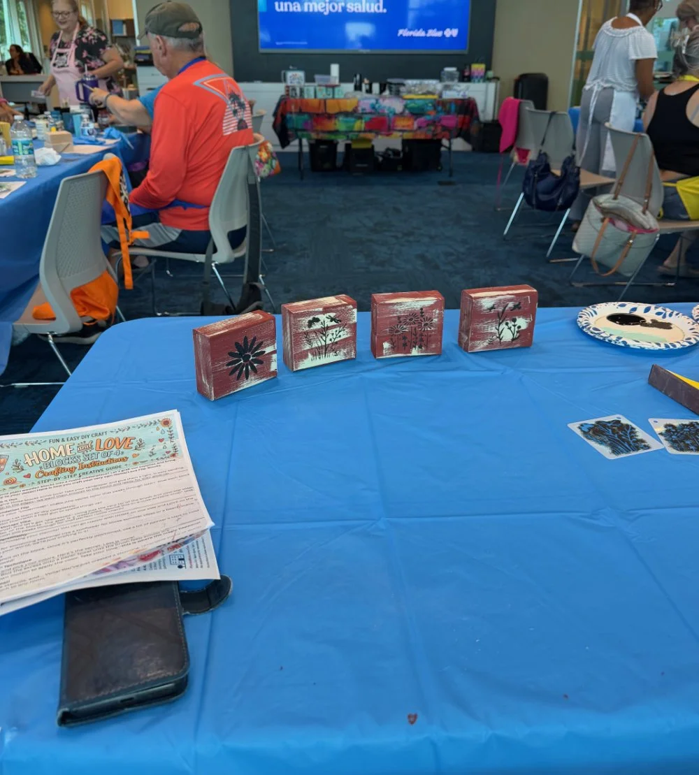 Decorative wooden blocks with floral patterns arranged on a blue tablecloth at a crafting event, with people seated at tables in the background.
