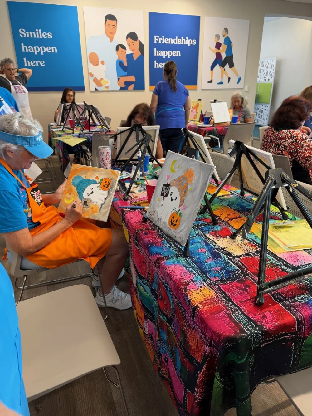 A group of elderly people painting Halloween-themed artwork at a craft event in a colorful room with wall posters that say 'Smiles happen here,' 'Friendships happen here,' and illustrations of people exercising.