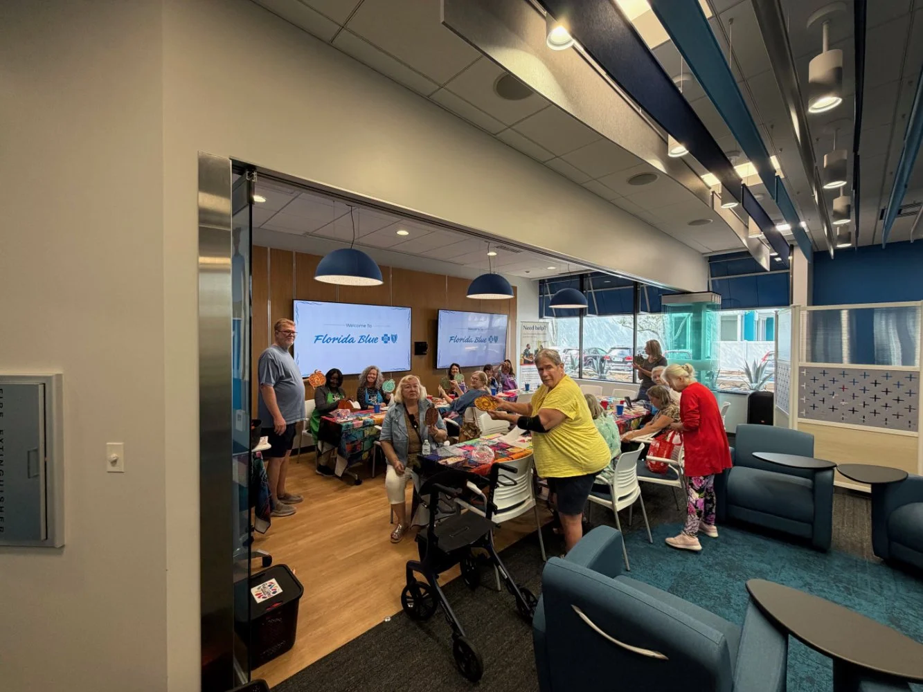 Group of people sitting and standing around tables in a room with two large screens displaying 'Welcome to Florida Blue' during a social gathering or event.