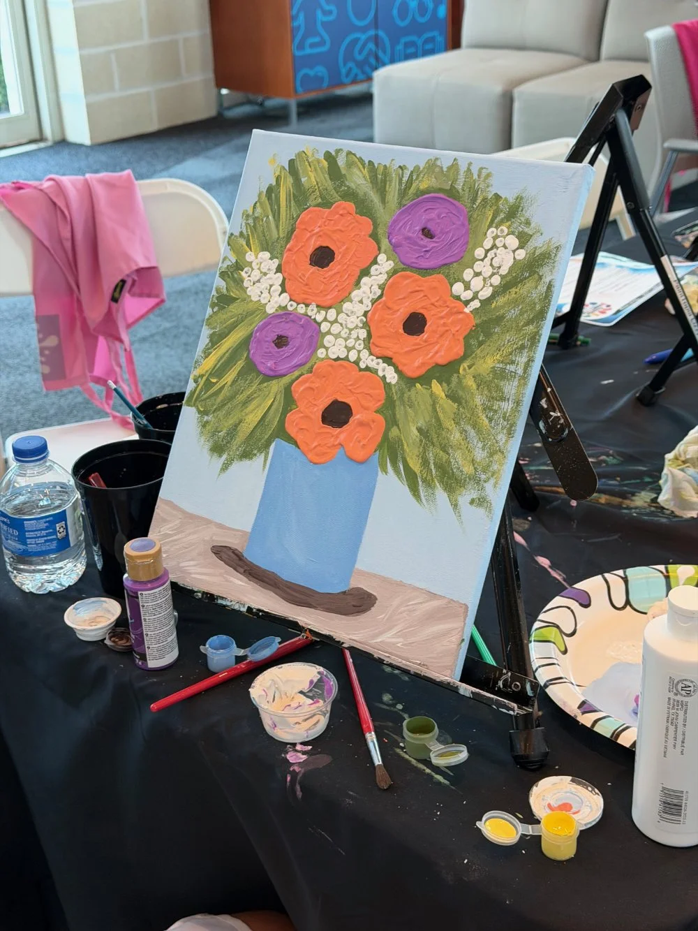 A colorful painting of a bouquet of flowers in a light blue vase, featuring large orange and purple flowers and white accents, on an easel at an indoor art station.