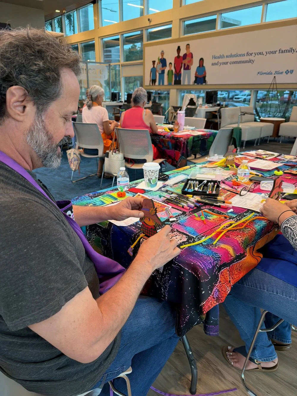 A man with gray hair and a beard sitting at a colorful table, engaging in a arts and crafts activity with paper cutouts and various art supplies. In the background, a few women are seated at the same table, and a large window with a banner above feat