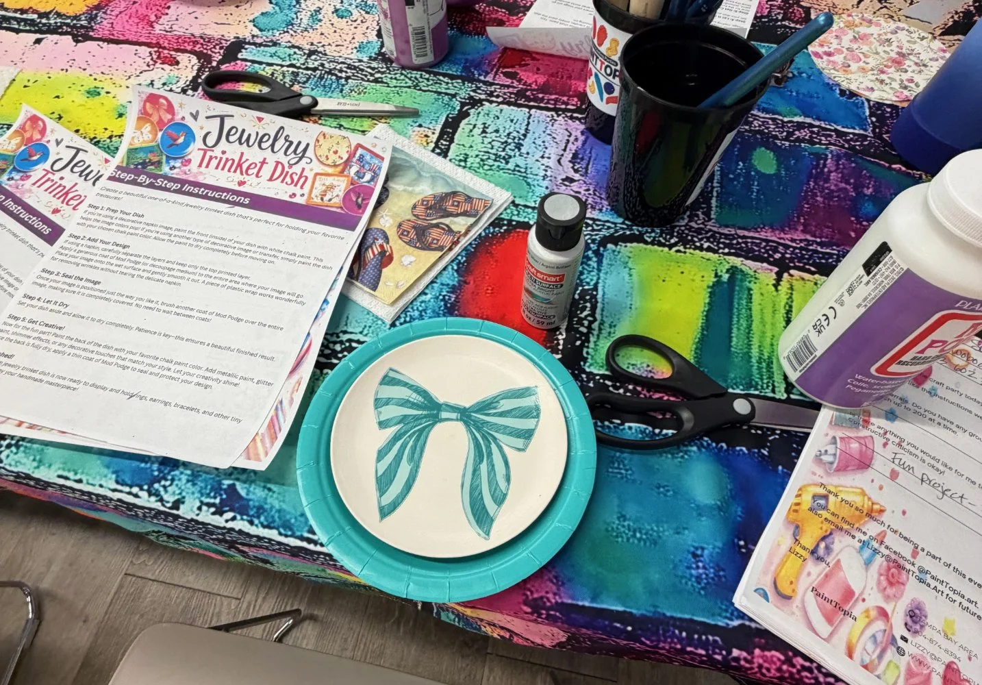 A colorful table with crafting supplies, including scissors, glue, a paper plate with a blue striped bow, paint bottles, and instruction sheets for making a jewelry trinket dish.