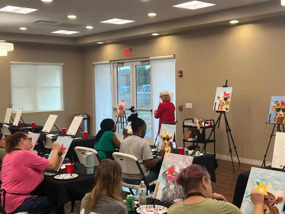 Participants attending a painting class in a large room, each sitting at a table with painting supplies, creating pictures of dogs wearing sunglasses with floral accessories. An instructor stands at the front, guiding the class, with completed painti