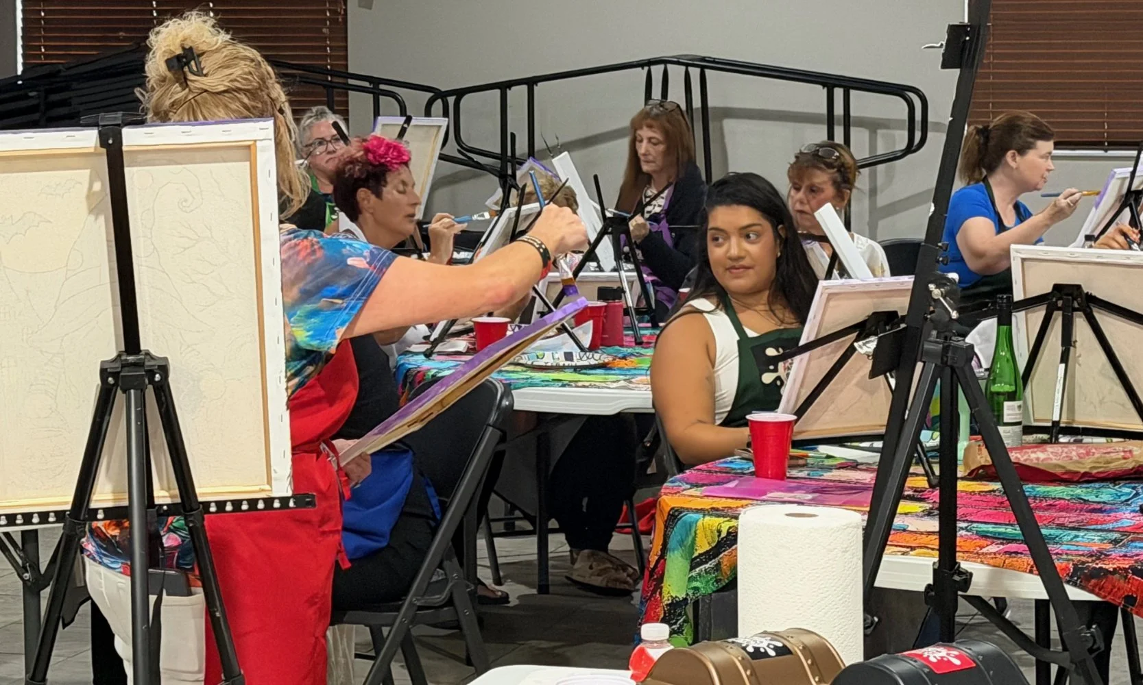 Group of people painting on canvases at an indoor art class. Some are focused on their work, others are looking at their canvases. The tables are covered with colorful tablecloths, and painting supplies are visible.
