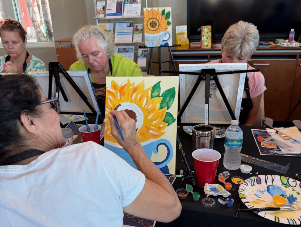 Group of elderly women painting sunflower pictures on canvases during an art class.