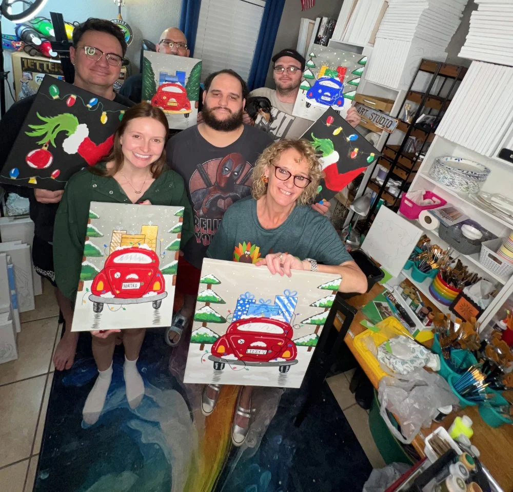 Group of six people in an art studio displaying holiday-themed paintings featuring red cars and Christmas decorations.