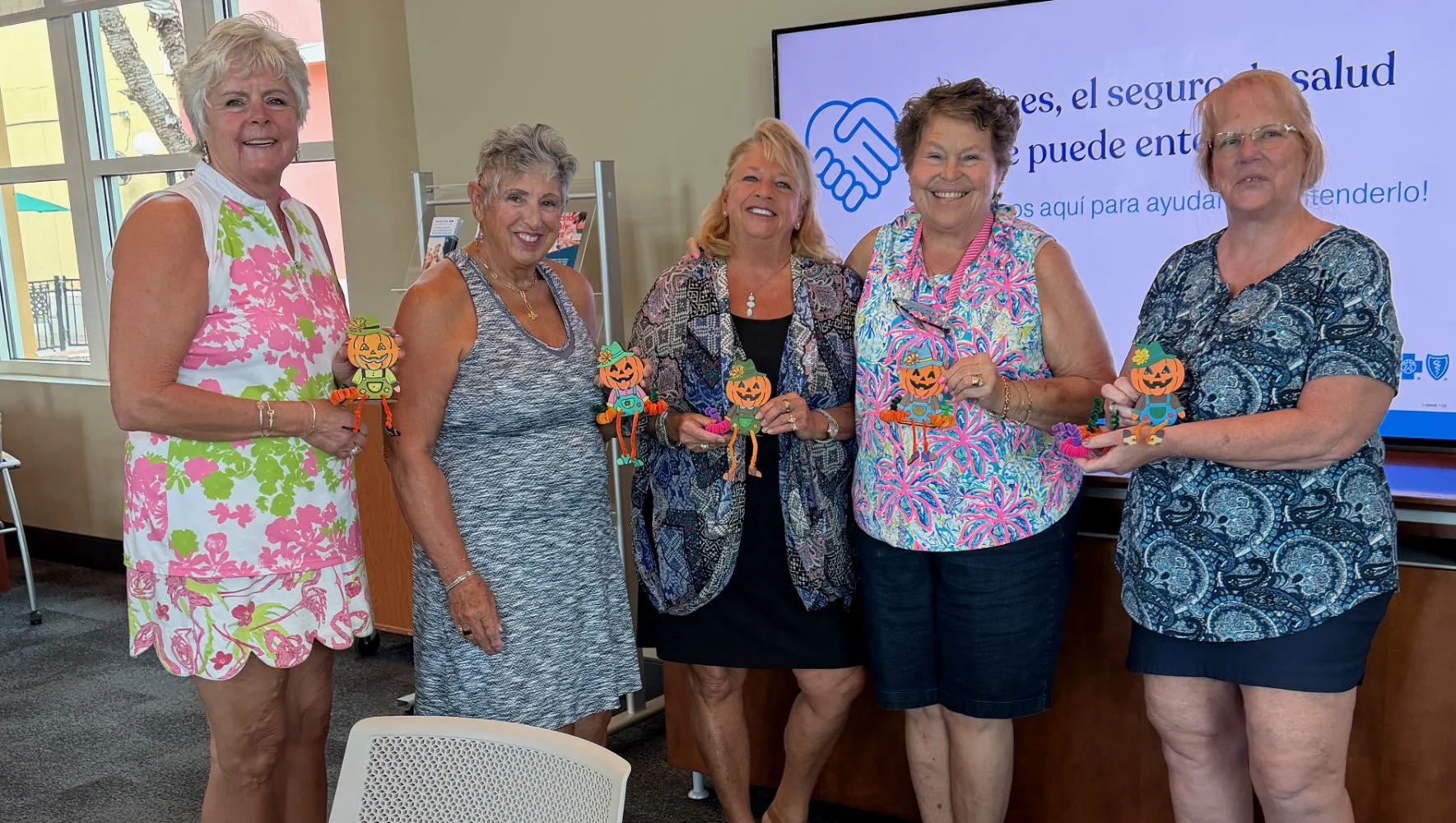 Five women standing together indoors, smiling, holding pumpkin and scarecrow decorations, with a large screen behind them displaying Spanish text and a handshake icon, suggesting a community or educational event.