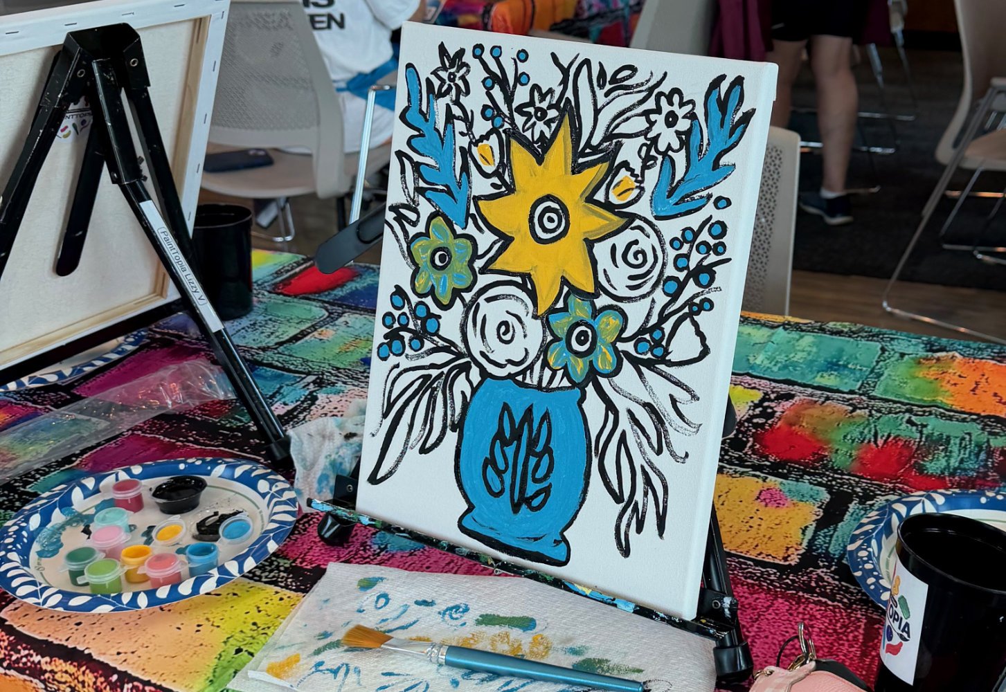 A colorful painting of a yellow star-shaped flower and smaller blue flowers in a blue vase on canvas, with black and white outlines and some painted details, on a table with art supplies at an art event.