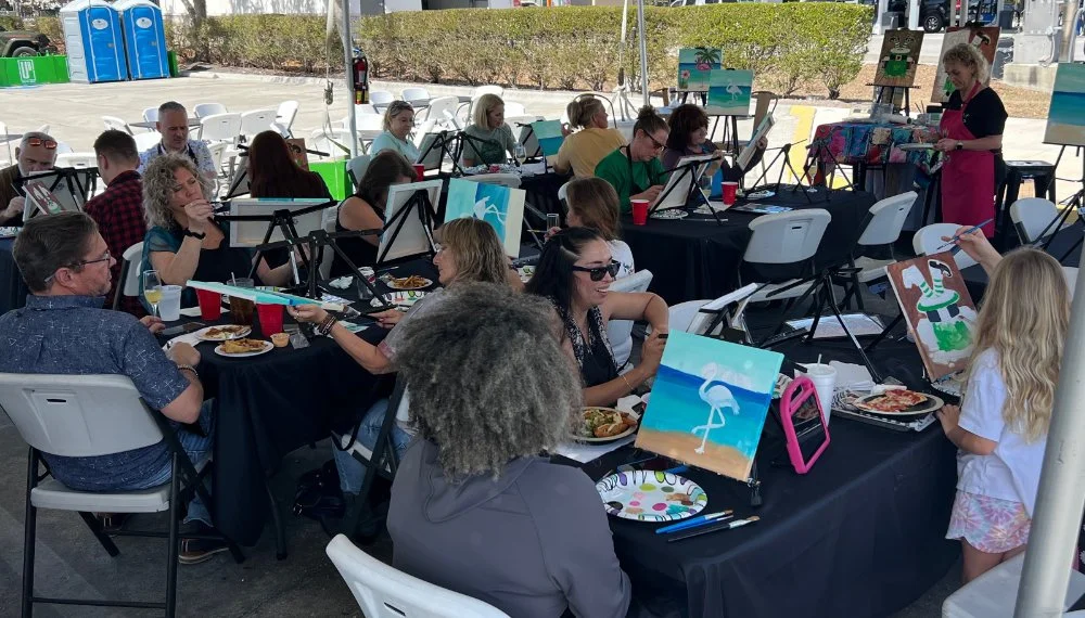 People participating in a group painting event outdoors, seated at tables with art supplies, with an instructor standing nearby.