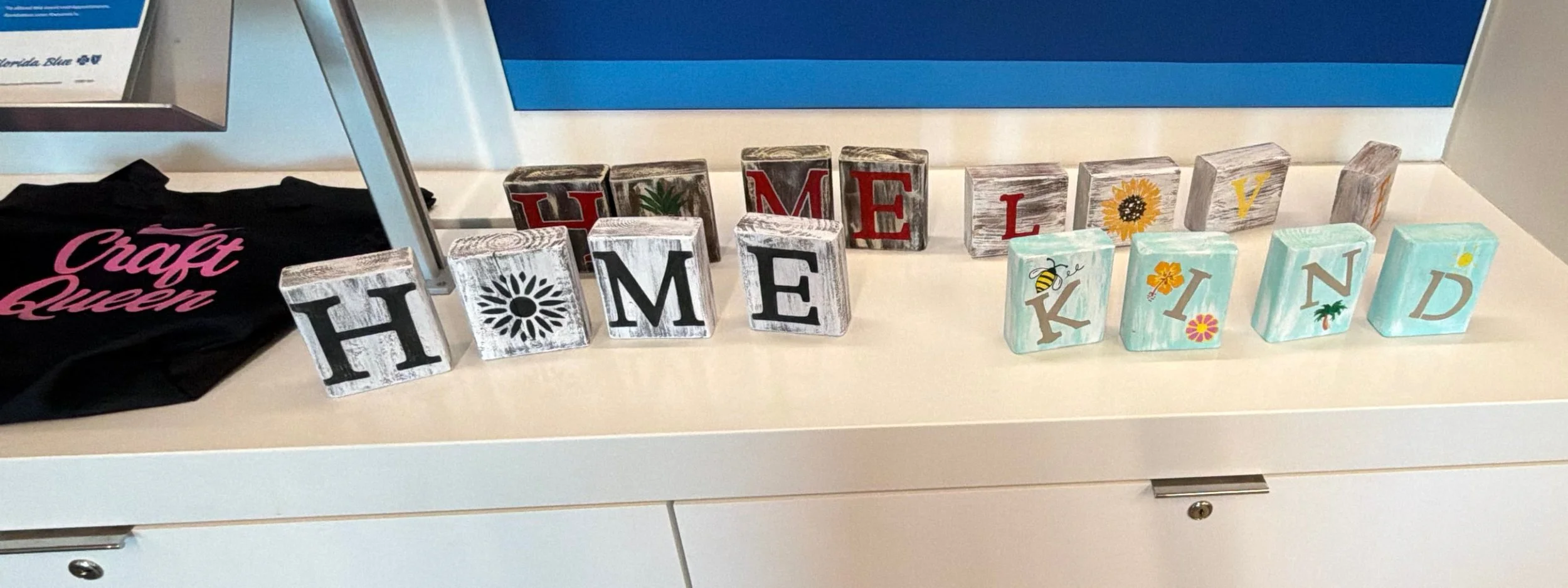 Decorative wooden blocks spelling out "HOME" and "KIND" with colorful designs and images, arranged on a white countertop in front of a blue background.