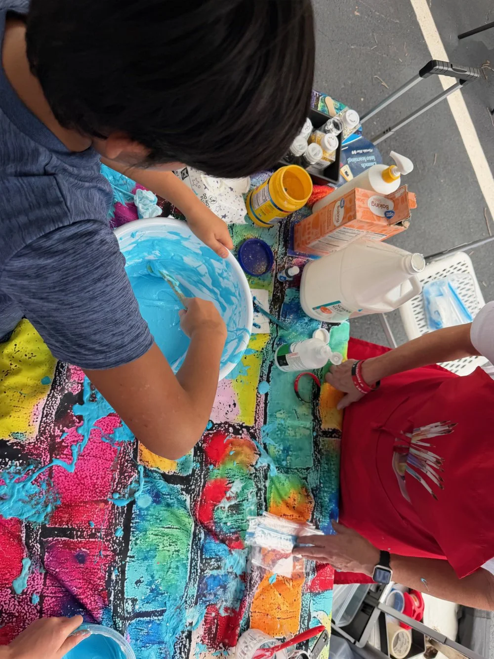 Children creating colorful slime on a table covered with vibrant paint splatters at an outdoor event.