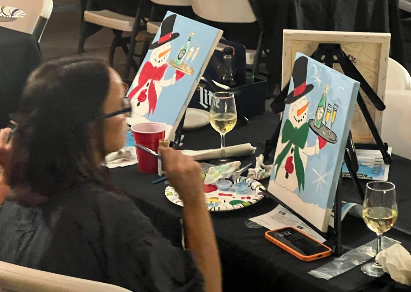 A woman sitting at a table painting holiday-themed snowman artwork, with two snowman paintings on easels, glasses of wine, and painting supplies on the table.
