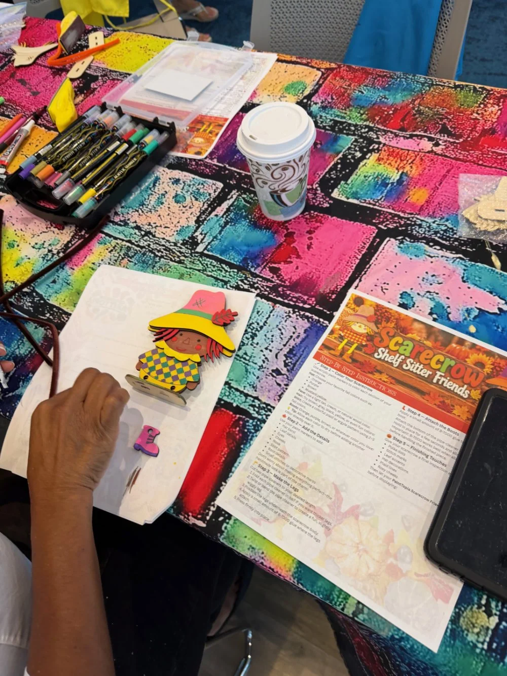 Colorful table with arts and crafts supplies, a decorated paper cup, a paper craft of a scarecrow, and a flyer titled 'Scarecrow Shelf Sitter Friends' at an arts and crafts event.