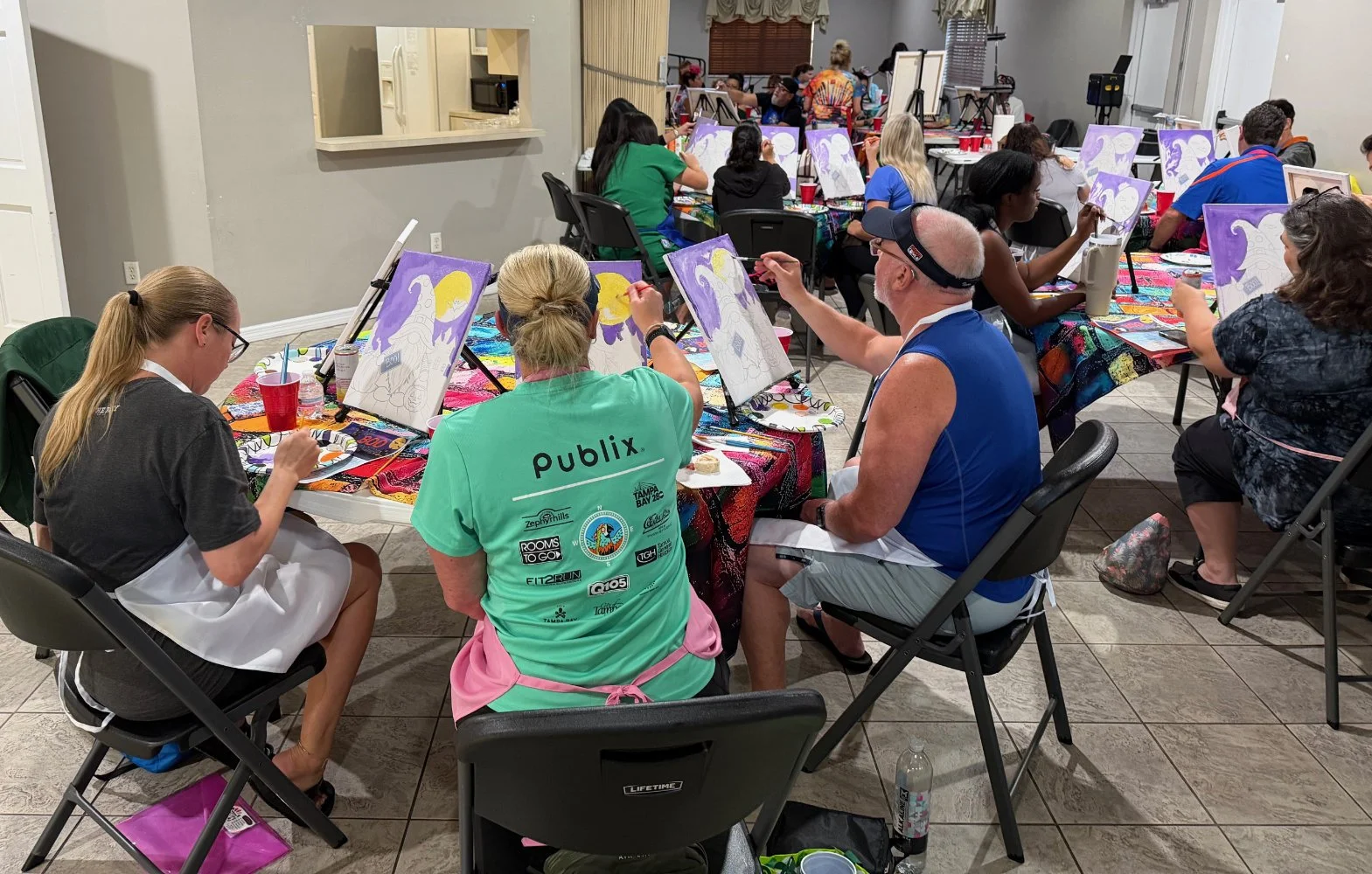 People participating in a group painting class, sitting at tables with canvases, paintings of a mermaid and moon, art supplies, and colorful tablecloths in a well-lit indoor space.