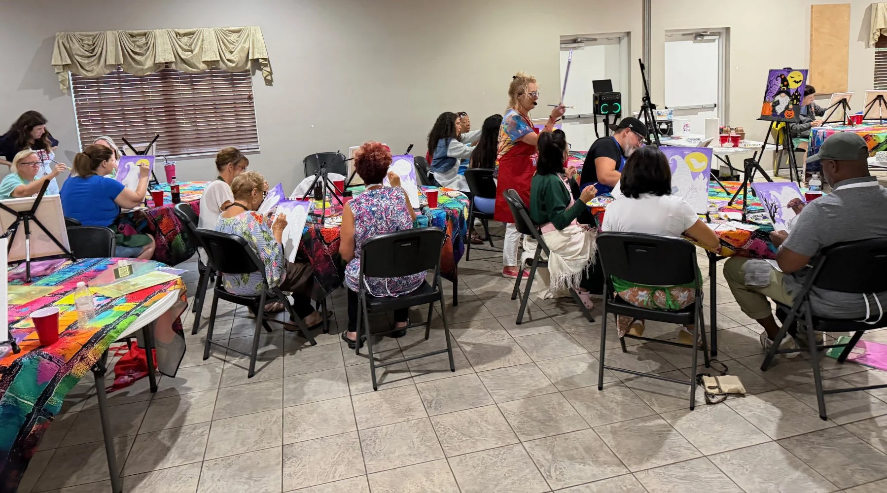 People participating in a watercolor painting class in a room with colorful tablecloths, painting easels, and art supplies, with a teacher giving instructions.