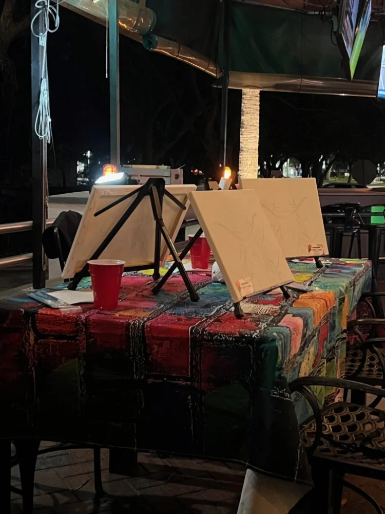 An outdoor painting setup on a table covered with a colorful cloth, with blank canvases on small easels, a red cup, and painting supplies, at night.