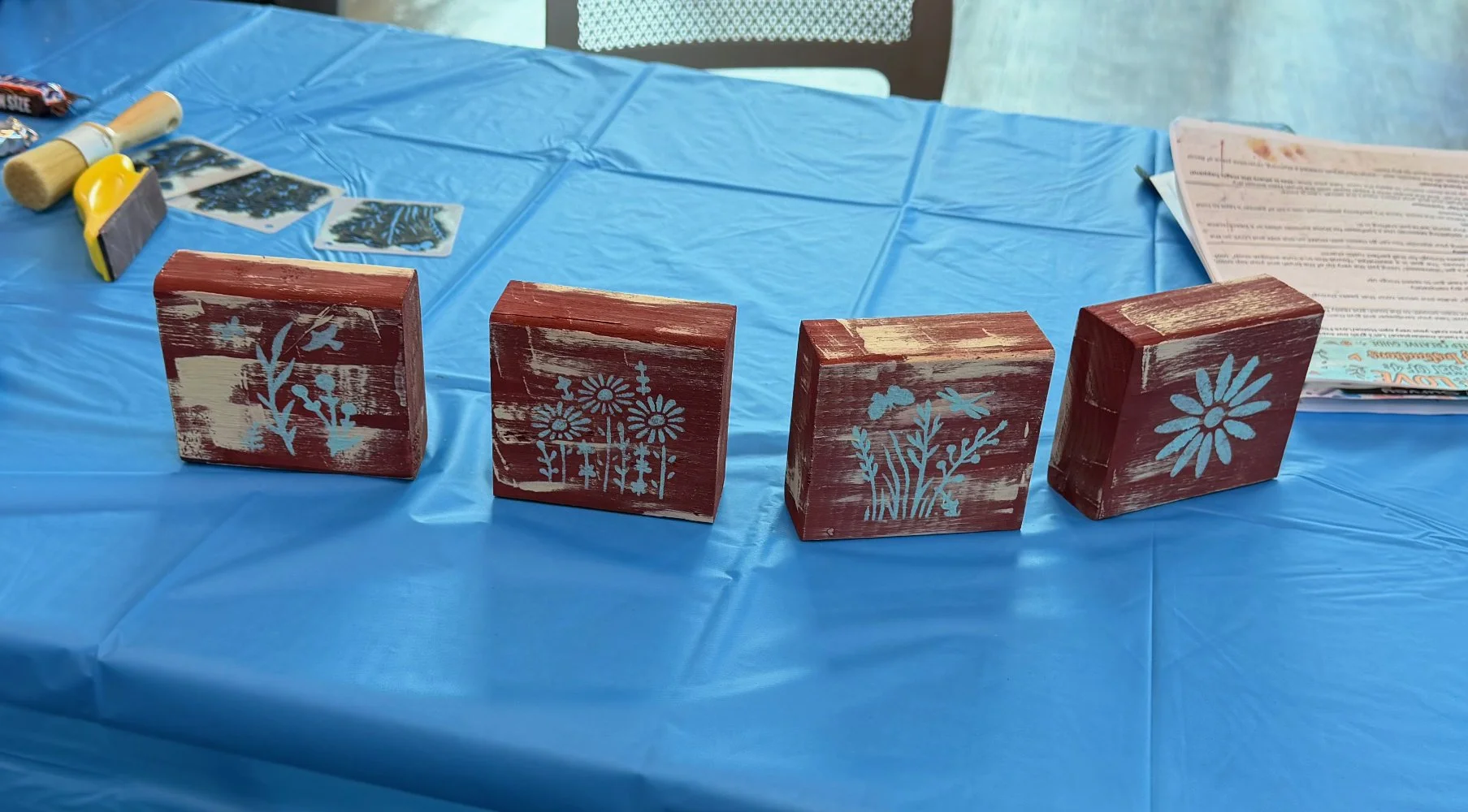 Four wooden blocks painted red with white distressed finish, each with a different floral stencil design in light blue, placed on a blue plastic-covered table. In the background, there are paint supplies, a printed instruction sheet, and some small s