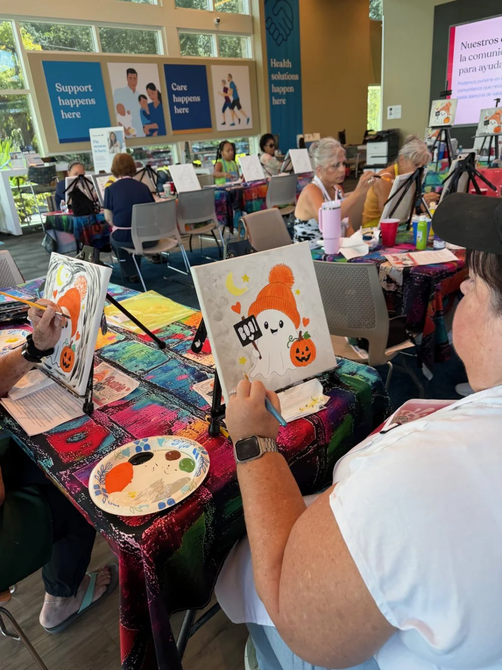 Older women painting Halloween-themed pictures of a ghost, pumpkin, moon, and stars at a table covered with a colorful tablecloth in a bright indoor setting during a group art activity.