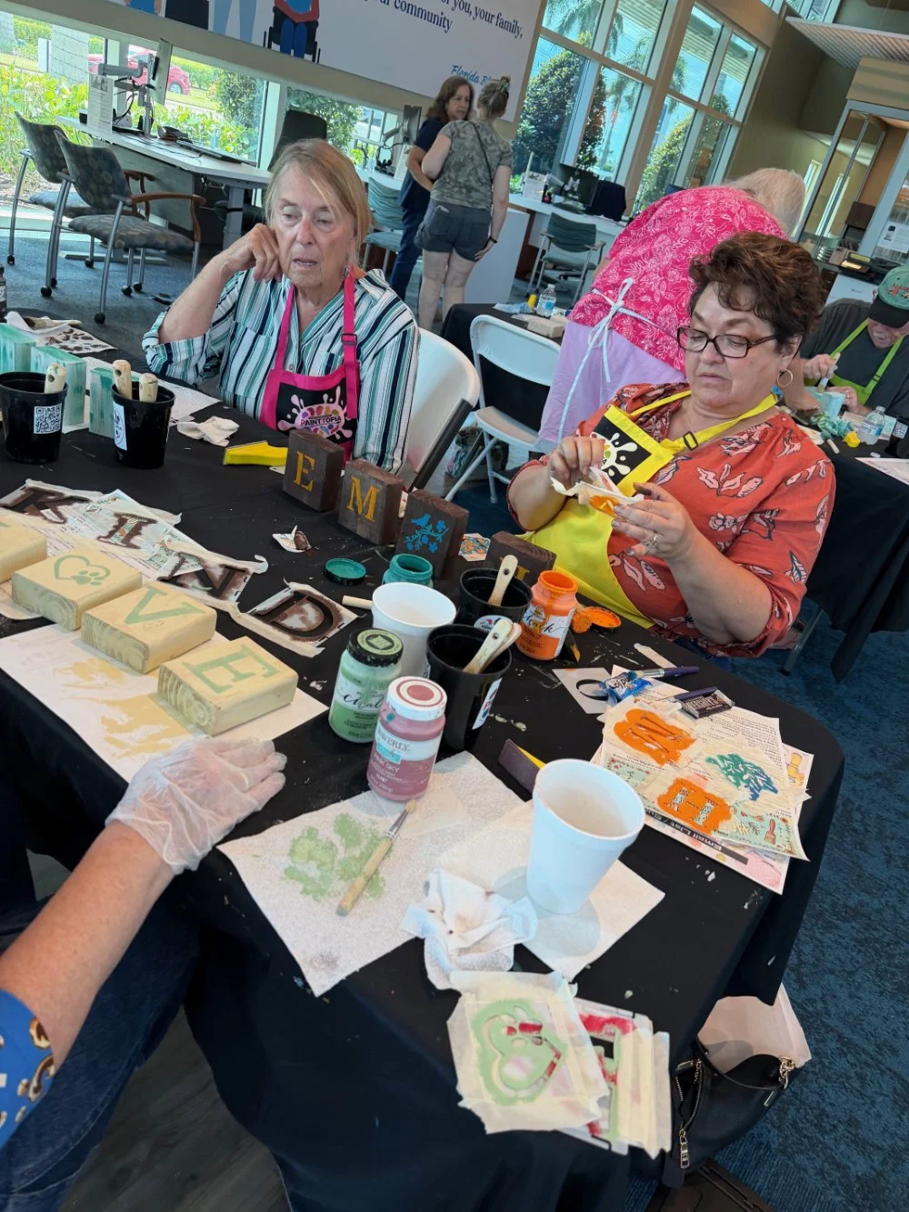 People participating in a craft workshop, painting and decorating wooden blocks with letters and designs at a table in a bright indoor space.