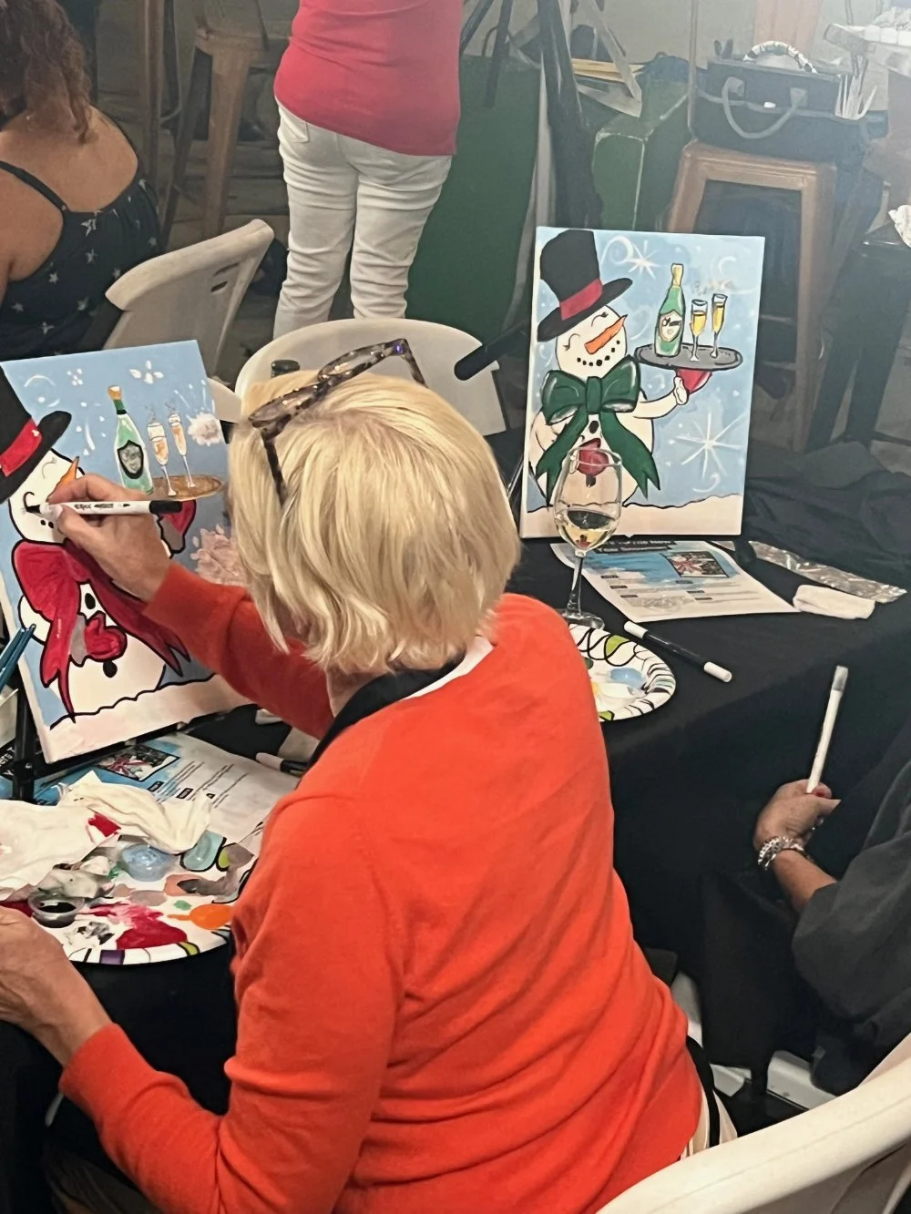 A woman with blonde hair, wearing an orange/red sweater, is painting a holiday-themed snowman on a canvas. The snowman has a black top hat with a red band, an orange carrot nose, and a green scarf, and is holding a tray with bottles and glasses. Ther