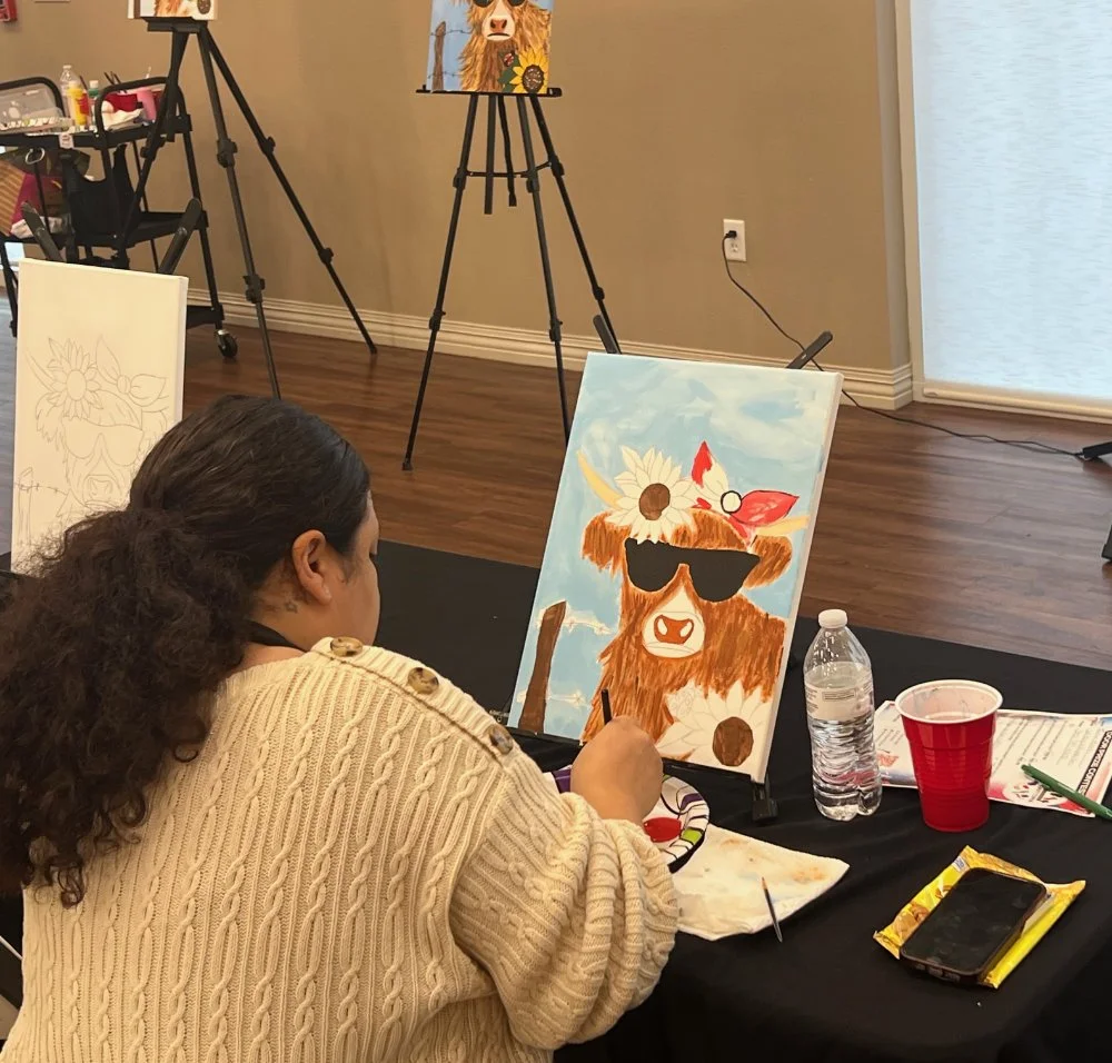 A woman sitting at a table painting a cartoon-style cow with a flower crown and sunglasses. The painting features a blue sky background with clouds. There are art supplies, a water bottle, a red cup, and a phone on the table. Other canvases and art e
