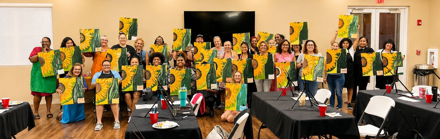 Group of smiling women holding up sunflower-themed paintings in a room with tables and chairs, celebrating art or a craft event.