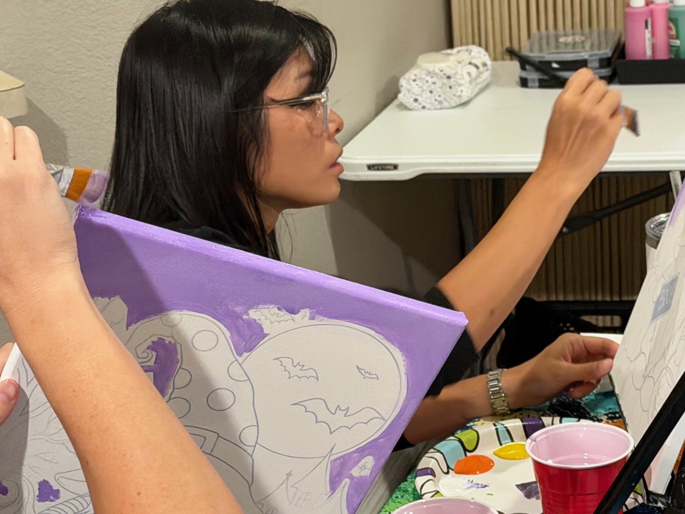A woman with black hair and glasses painting Halloween-themed artwork with bats and a moon on a canvas. There are art supplies, including paints and cups, on the table.