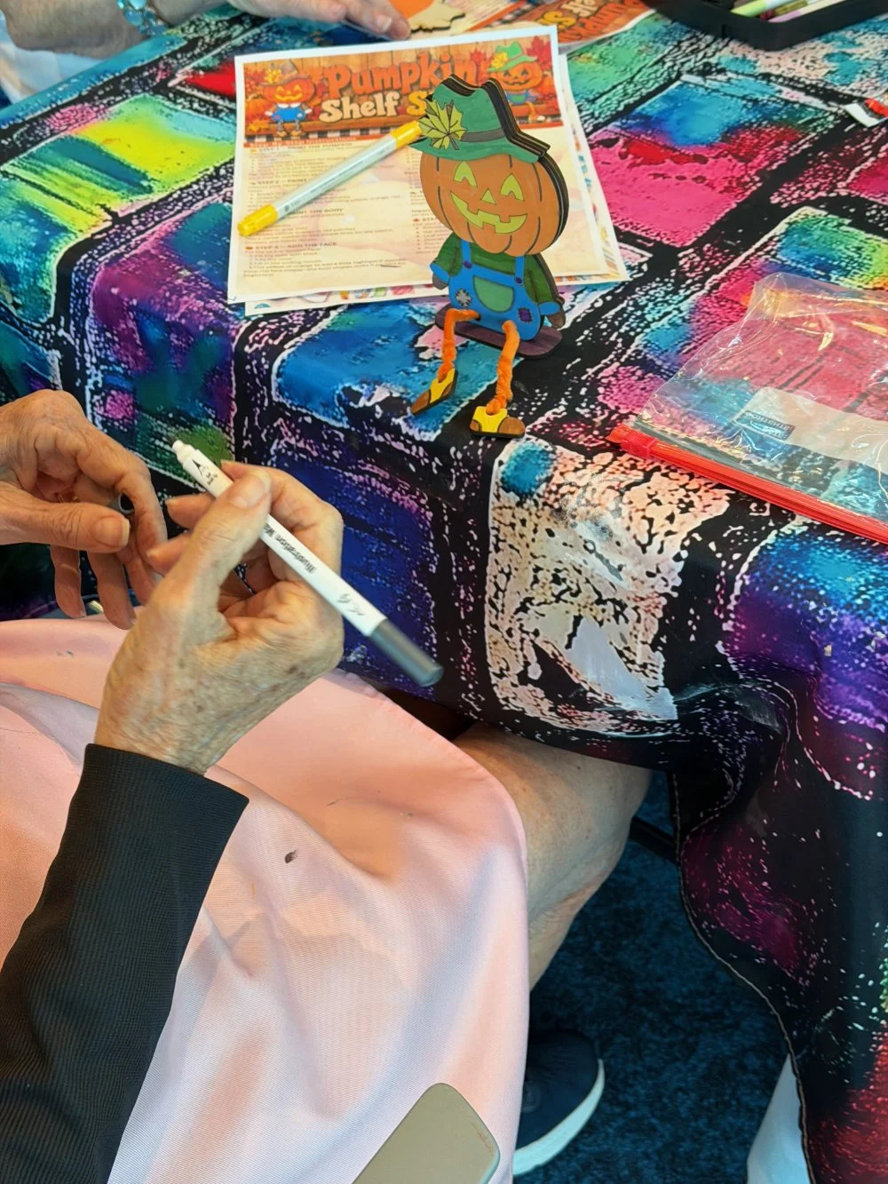 A colorful table with a pumpkin-themed decoration, a menu, and a pen. The tablecloth has a vibrant, abstract pattern. A hand is visible holding a marker, and part of a person wearing a pink skirt and black shoes. The pumpkin decoration has a cartoon 