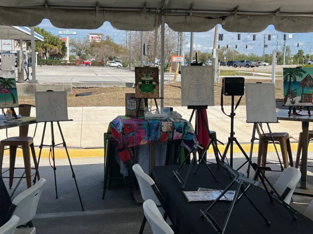 An art booth under a tent with various paintings and sketching supplies, with a view of a parking lot and traffic lights outside.