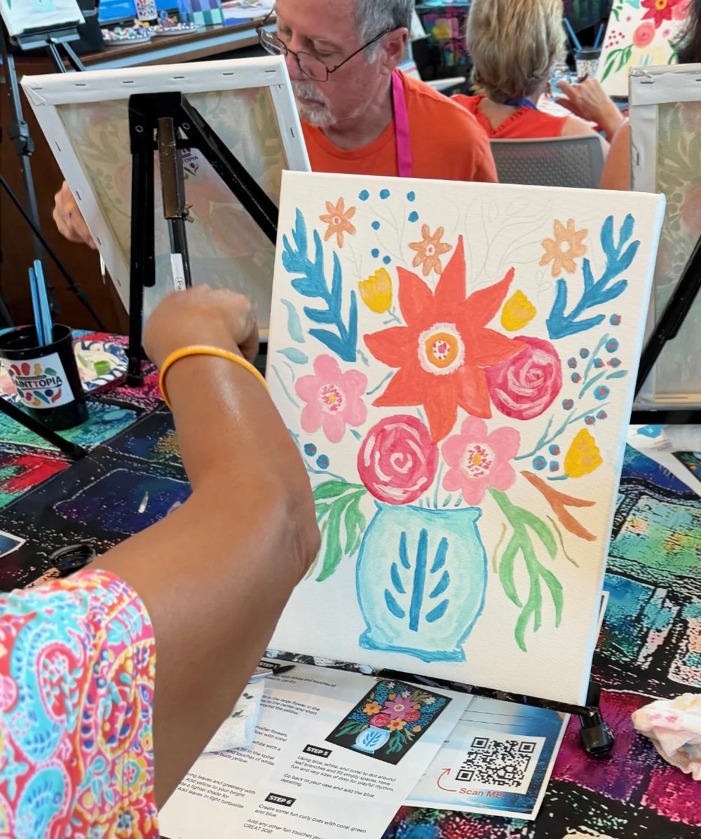 A person is painting a colorful floral picture on a canvas at an art activity event. The painting features bright flowers in a blue vase. In the background, other participants are also engaged in art projects.