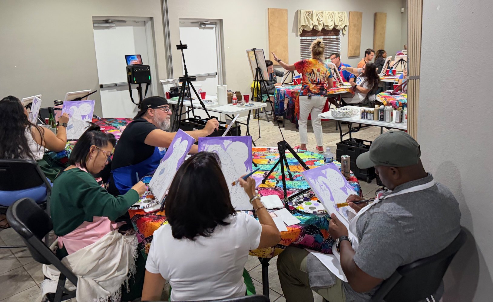 People in a painting class, some are painting on their canvases, while an instructor guides a participant standing at the front of the room. Art supplies and colorful tablecloths are visible.