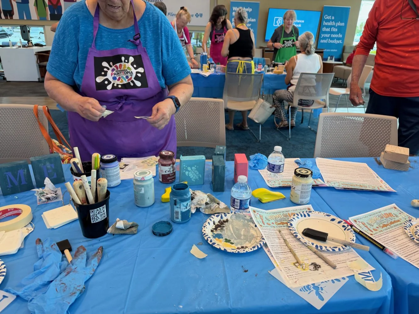 A woman wearing a purple apron standing at a table with art supplies, including paint, brushes, and paper, during a painting class at Paintopia. Other people are in the background, some sitting and some standing, with materials and advertisements vis