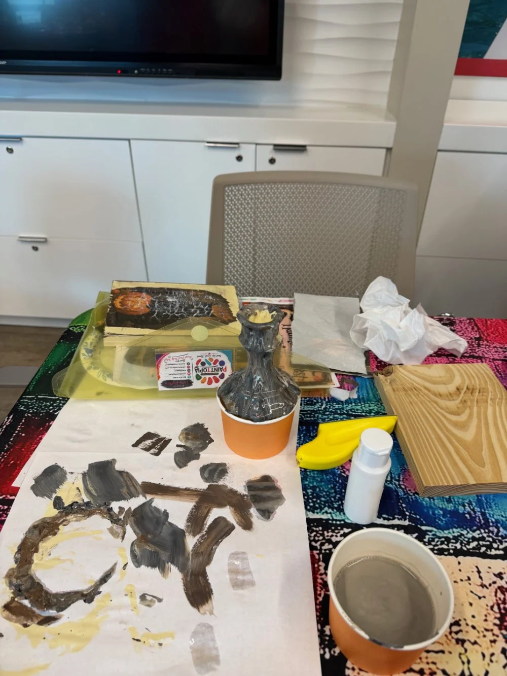 A cluttered table with a painted paper, a small ceramic vase, a banana, a white pill bottle, a cup of coffee, tissue paper, a wooden board, and a box of paint supplies. In the background, a chair, a closed cabinet, and a television are visible.