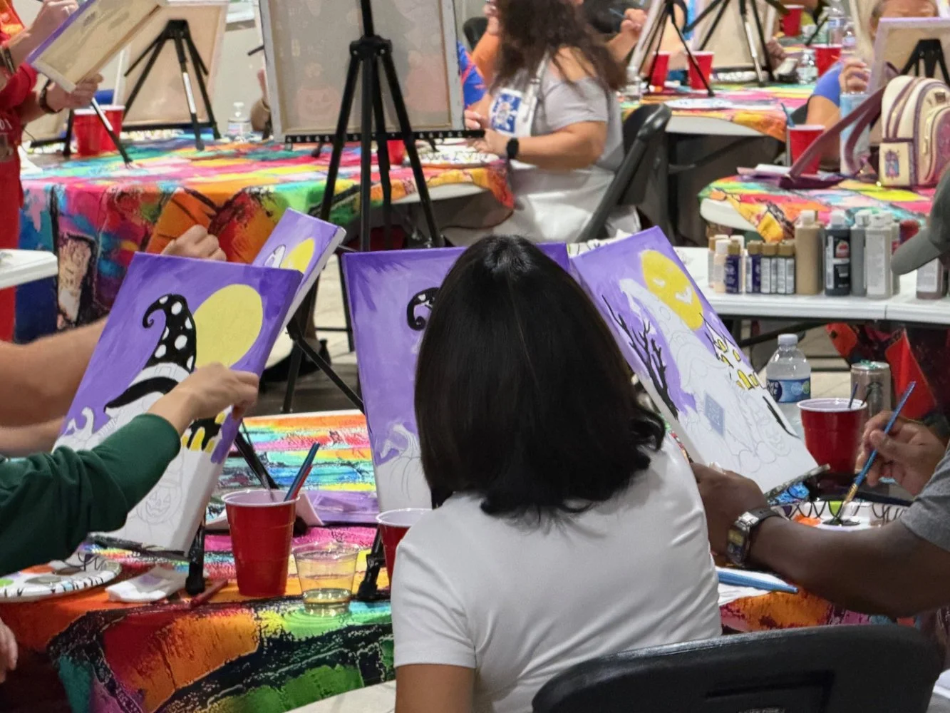 People painting Halloween-themed pictures of a white ghost with a yellow moon in the background, seated at a table covered with a colorful, rainbow-patterned cloth at a group art event.