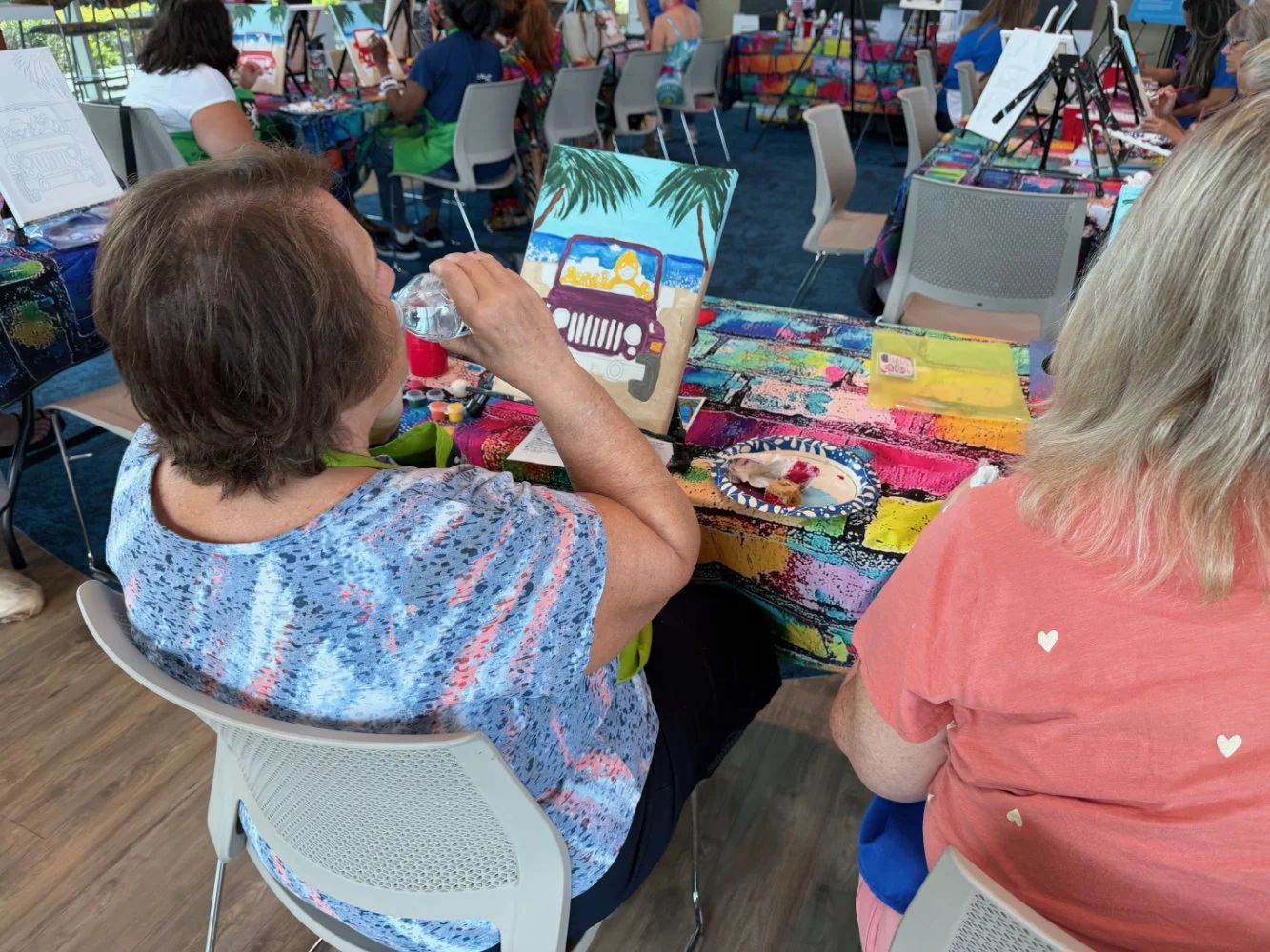 Two women sitting at a colorful, splattered paint table on a boat or indoor pavilion, completing a painting of a beach scene with a car, palm trees, and water. Other people are visible in the background also painting.