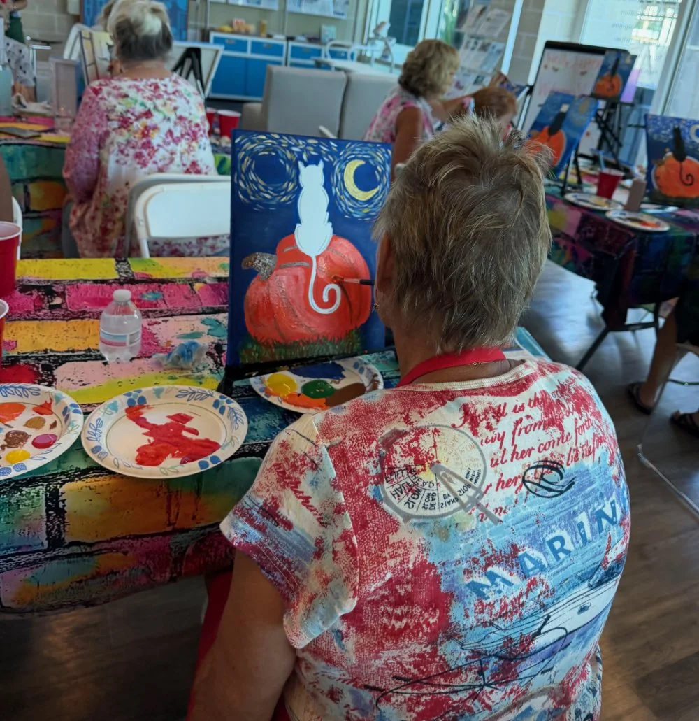 Older woman painting a canvas with a fall-themed scene featuring a white cat, a pumpkin, and a night sky with a crescent moon.
